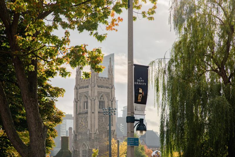 photo of U of T Soldier's Tower in the background. Two large, green trees on the side and a blue banner reading "Defy Gravity" in the foreground. 