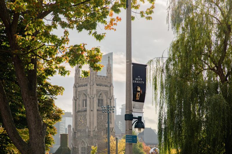 photo of U of T Soldier's Tower in the background. Two large, green trees on the side and a blue banner reading "Defy Gravity" in the foreground.