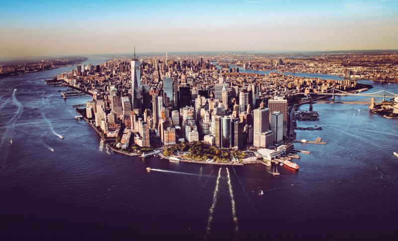 An aerial photo of Manhattan, New York City. You can see the iconic skyscrapers, the Hudson and East Rivers, and landmarks such as the One World Trade Center in the foreground and the Brooklyn and Manhattan Bridges in the distance.