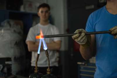 A student in safety glasses and gloves holds a steel tube in the flame of two welding torches until it glows red hot.