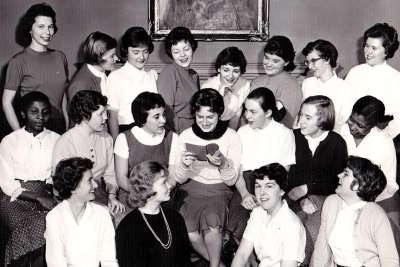 A group of young women in the fashions of 1958 sit and laugh together.