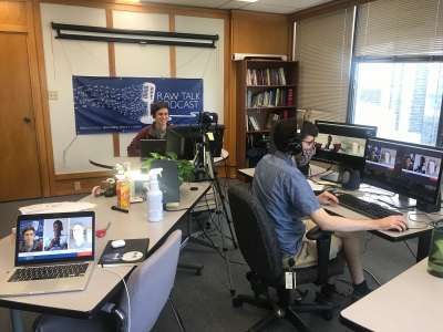 Jesse Knight sits in front of a film camera while Alex Jacob, wearing headphones, operates a bank of computers.