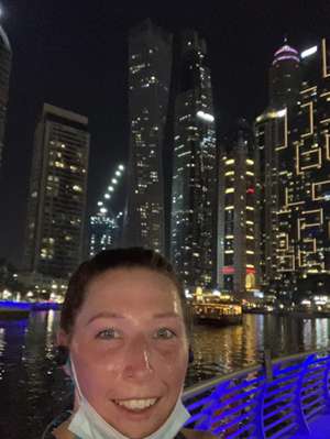 Brittney Scott pulls down her mask to smile, standing by a waterfront at night with lit-up skyscrapers across the water.