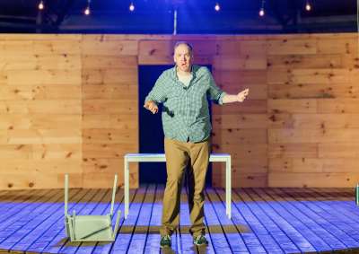 Mark Crawford on a stage with one overturned chair, looking exaggeratedly concerned.