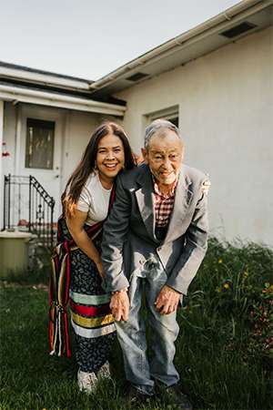 Angelique Belcourt and Fernand Belcourt laugh together in a garden outside a house.