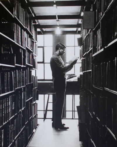 A black-and-white old photo of a young man standing between library shelves,