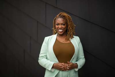 Desiree Kaunda-Wint smiles, standing by a building wall.