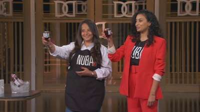 Two women stand on the Dragon's Den TV set, raising glasses. The glasses and their t-shirts have the word Nuba on them.