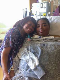 Carisse Samuel lies in a hospital bed and her mom, Bernadine, holds her hand and leans their heads together.