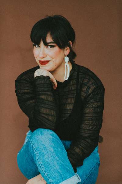 Riley’s headshot - Photo of Riley Yesno sitting in front of a light brown background. Riley has dark, short hair and is wearing white drop earrings, light blue jeans and a black long sleeve top.