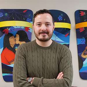 Jeff Ansloos smiles, standing in front of Indigenous artwork depicting people, land and flowers.