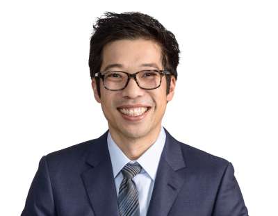 Headshot of Mark Tan smiling - Mark is a man with short, dark hair and wears glasses. He appears in front of a white background and is wearing a dark blue suit with a light blue shirt and striped tie.  