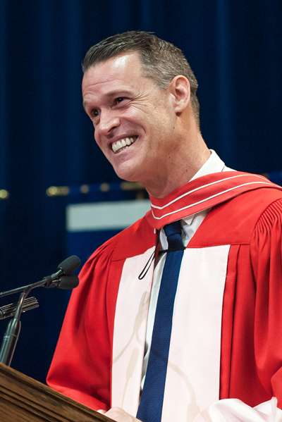 Mark Tewksbury laughs as he gives a talk at a podium. He is wearing academic robes.