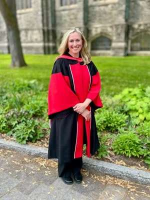 A person wearing a convocation gown and posing outdoors.