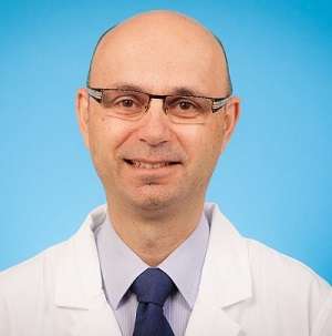 Michael Cusimano wears a lab coat and stands in front of a blue wall, smiling