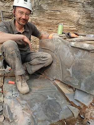 Jean-Bernard Caron crouches beside a rock, on which a long ridge between two flat areas marks the shape of a fossil.