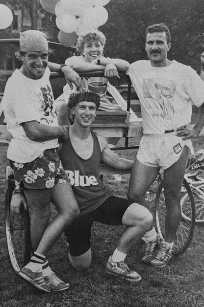 In a old photo, three young men in shorts smile as they lean on a cart decorated with balloons.