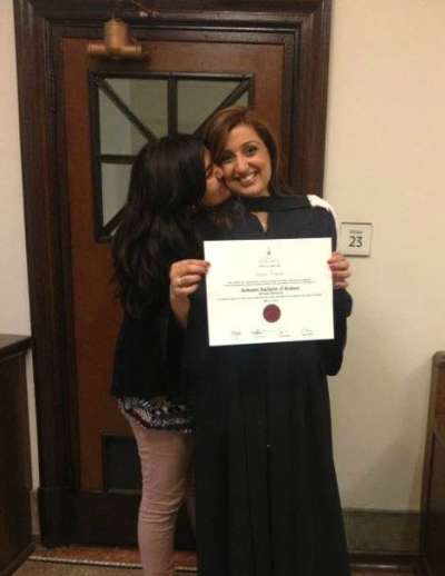Samra Zafar, in academic robes, holds up her framed degree while her teen daughter kisses her cheek.
