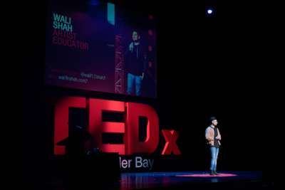 Walk Shah stands alone on an enormous stage with the TEDX logo behind him.