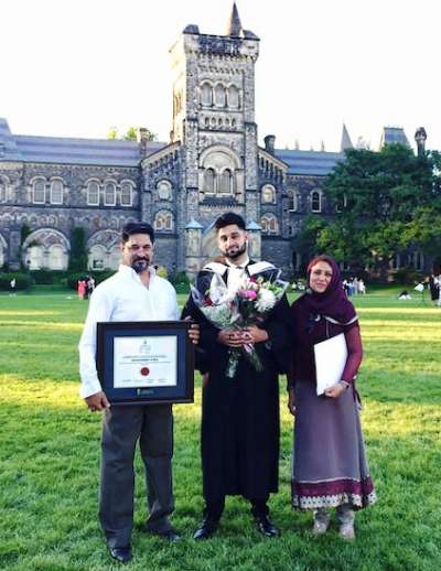 Wali Shah, in academic robes, stands with his parents on Front Campus on a sunny evening. His dad holds his framed degree.