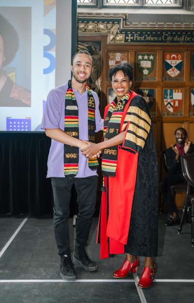 Etienne Oshinowo stands with Rhonda McEwen in Hart House, both wearing Black Graduation stoles.