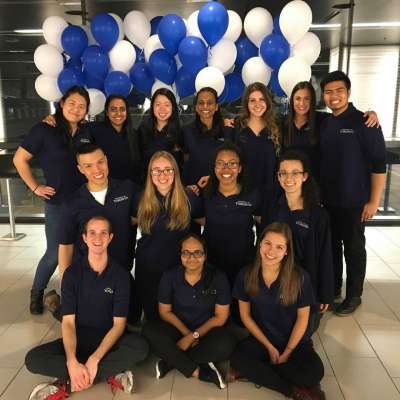 14 people wearing U of T t-shirts pose, smiling, in front of blue and white balloons.