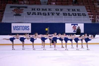 Women skaters in the U of T colours of blue and white sweep across the ice with legs lifted.