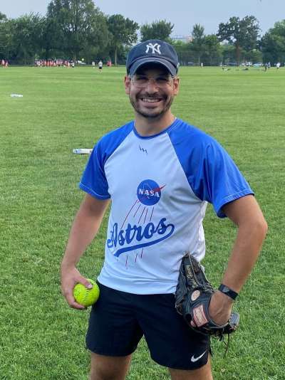 David Harary smiles while standing on a baseball outfield, holding a glove and ball and wearing a NASA Astros t-shirt.