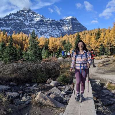 Lulu Li smiles as she crosses a plank bridge. Behind her is a gorgeous snowy mountain and a larch forest in fall colours.