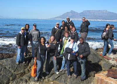 Marcus Singleton poses with a group on a rocky seashore. Across the water is Cape Town’s famous Table Mountain.