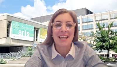 Melanie Woodin smiles as she chats on a video call. The virtual background behind her is a picture of Sidney Smith Hall.