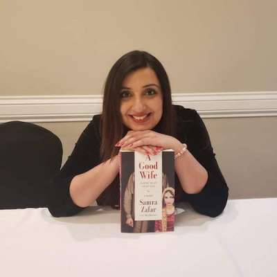 Samra Zafar smiles and holds her book. The cover says A Good Wife and the image is a faceless man looming over a young woman.