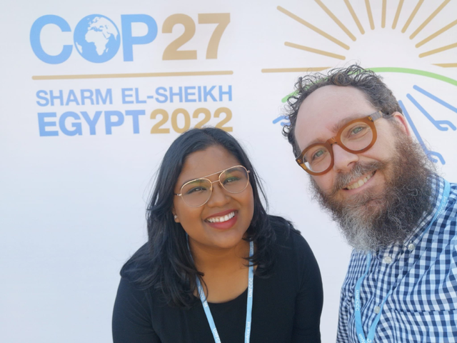 Anjum and a coworker stand in front of a placard, both smiling