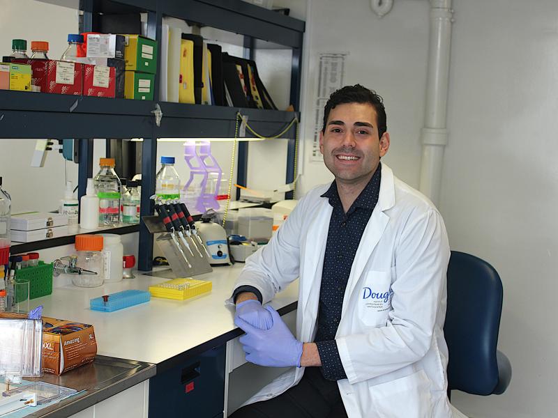 Daniel Almeida smiles as he sits at a lab bench crowded with scientific equipment