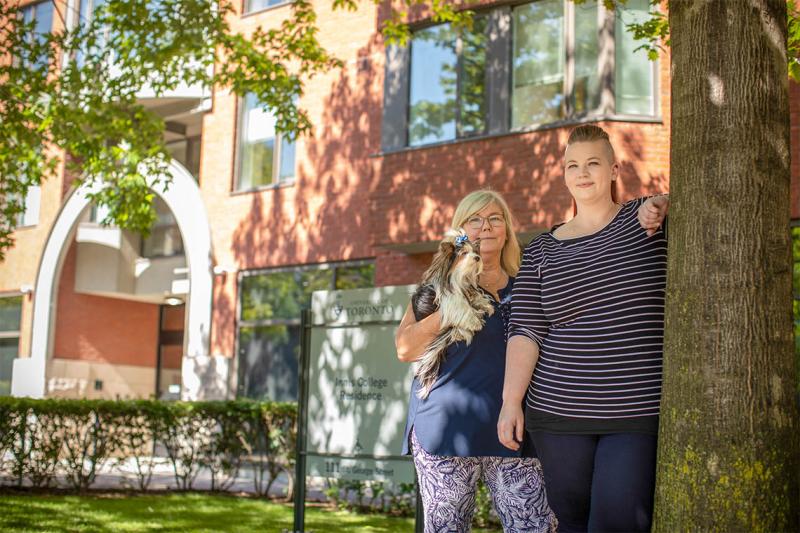Nicole McLeod and her mom stand under a tree outside Innis College, holding their tiny dog.