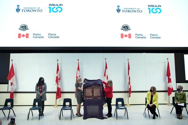 At a podium in a classroom, Christine Allen and Christine Loth-Brown lift a cloth to unveil a metal plaque.