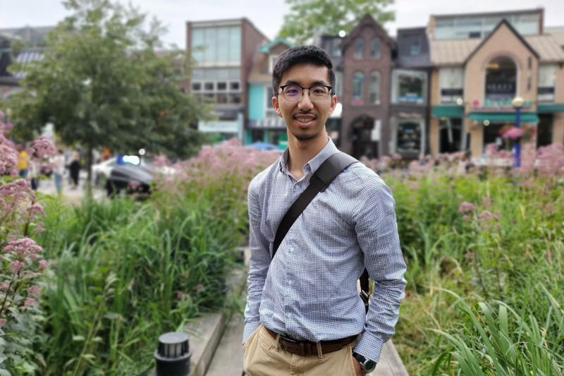 Lyndon Chan smiles while standing in a park beside a shopping street.