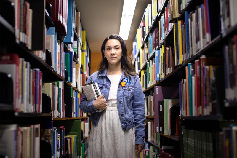 Mikayla Redden stands between tall library shelves, holding a stack of books and looking doubtful.