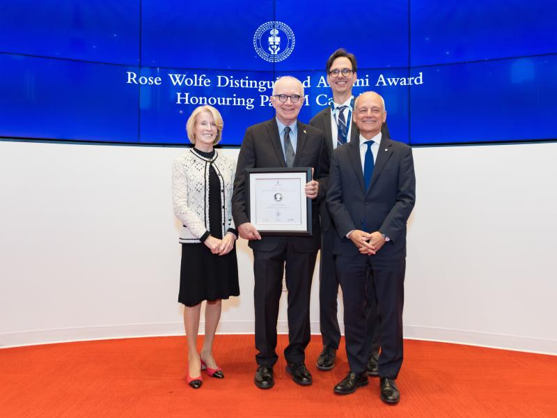 Paul Cadario stands on stage between Rose Patten, Corwin Cambray and Meric Gertler, holding his award and smiling