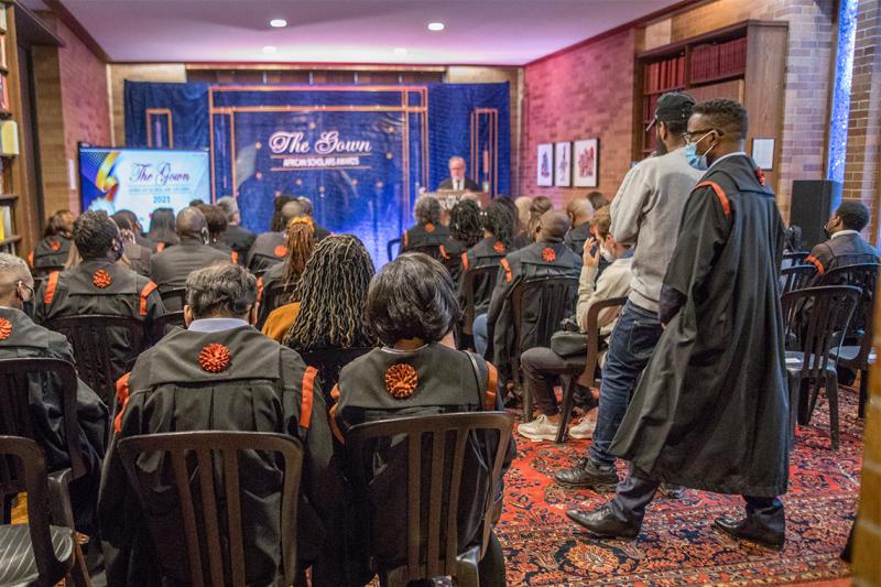 A group of people in masks sit in rows in a library room. They wear academic gowns with orange decorations.