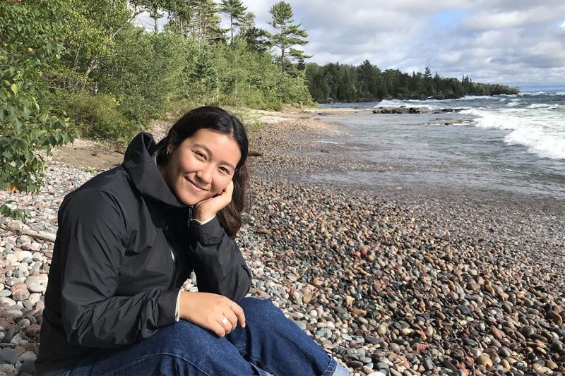 Alexandra Gaspar sits on a pebbled, tree-lined beach, leaning her head on one hand and smiling. Waves roll in behind her.