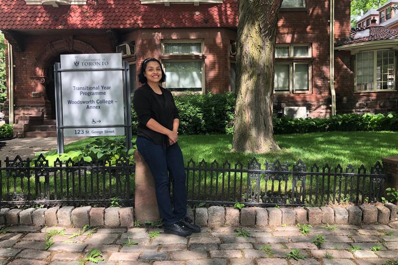 Ana Chiguichon smiles, standing on the sidewalk in front of a sign that reads, Transitional Year Programme.