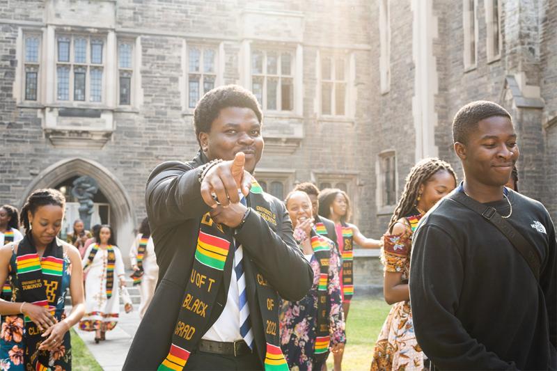 A young man smiles and points in celebration. He is wearing a stole with text on it: U of T Black Grad.