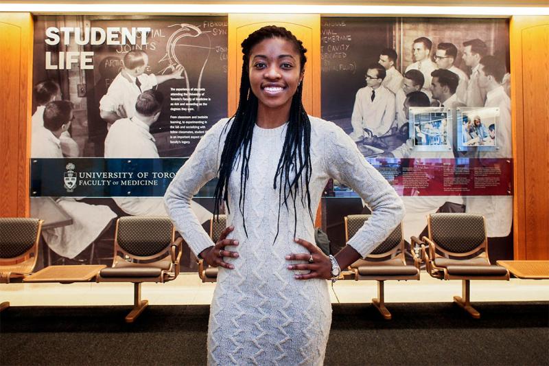 Chika Oriuwa smiles happily, standing in front of a display about the history of Student Life at the Faculty of Medicine.
