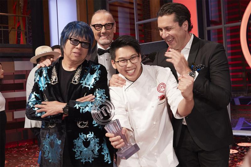 Christopher Siu laughs and holds up a plexiglass trophy shaped like the letter M while three men smile and hug him.