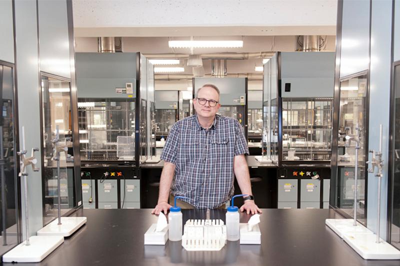 David McMillen leans on a steel bench in a large lab full of equipment contained in glass and plastic hoods.