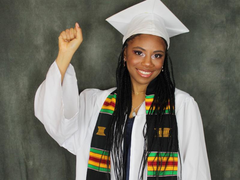 Denessia Blake-Hepburn raises her fist. She is wearing graduation robes and mortarboard hat, and a Black Grad kente scarf.