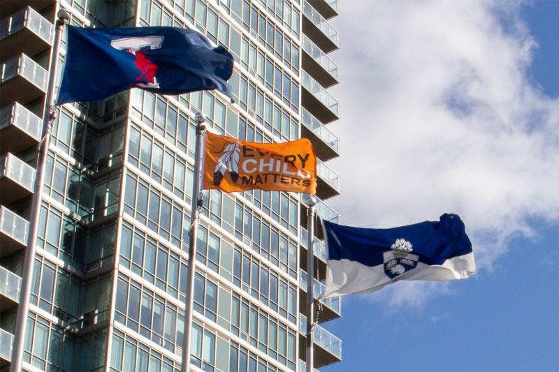 An orange flag with the words every child matters flies on a flagpole beside the U of T flag.