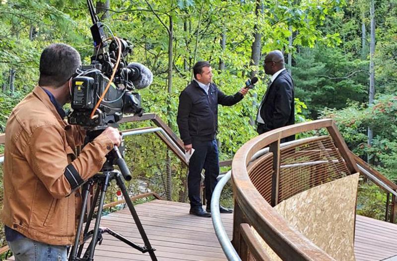 Wisdom Tettey chats with a CTV reporter on a curved wooden bridge, while a man points a large TV video camera at them.