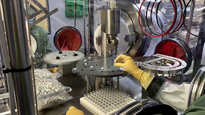 A worker reaches gloved hands through a portal into a sealed room, and places a tiny vial into a slot on a filling machine.